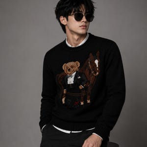 May include: A black long-sleeved knit sweater featuring a central graphic of a brown teddy bear wearing a black tuxedo and bow tie, standing next to a brown horse. The sweater has a ribbed crew neckline, cuffs, and hem, worn over a white collared shirt.