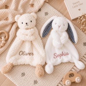 May include: A cream-colored plush bear security blanket embroidered with the name Oliver and a white plush rabbit security blanket with navy and white striped inner ears embroidered with the name Sophia, both featuring soft, textured fabric.
