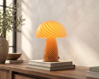 Op de afbeelding: Een paddestoelvormige tafellamp van oranje en wit gedraaid glas. De lamp heeft een afgeronde kap en een taps toelopende voet met een kleine metalen aan-uitknop aan de zijkant. De lamp is verlicht en staat op een stapel van twee boeken.