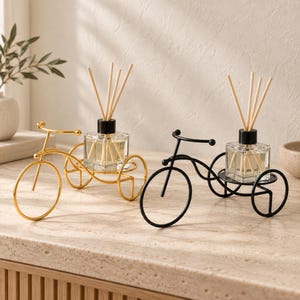 Puede incluir: Dos soportes de metal con forma de bicicleta, uno dorado y otro negro, cada uno con un frasco difusor de vidrio cuadrado con tapa negra y varias varillas de madera. Los marcos de las bicicletas cuentan con una rueda delantera y una plataforma.