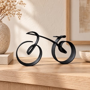 May include: A matte black metal sculpture shaped like a bicycle. The design features continuous, fluid lines forming the frame, two circular wheels, and a seat. The structure is constructed from a single piece of curved metal ribbon, creating a minimalist silhouette that stands upright on a flat surface.