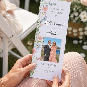 May include: A vertical wedding program card featuring a floral border with pink, blue, and white flowers. The text reads: Welcome to the wedding of Lill Susann and Martin, July 04, 2026, Grimsby Estate, Statham. It includes a photo of a couple.