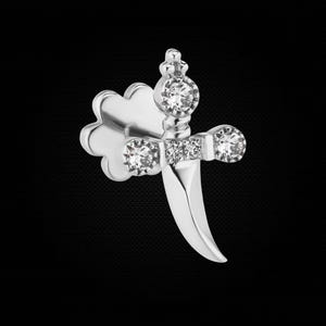 May include: A silver-toned stud earring shaped like a dagger with a curved blade. The hilt features three clear, round-cut stones set horizontally, and a larger clear stone is set at the pommel. A four-lobed flower-shaped backing is visible.