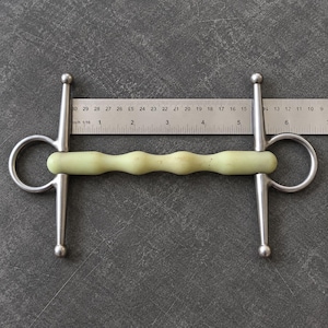 Pode incluir: Um bridão com embocadura texturizada em amarelo pálido e hastes laterais de aço inoxidável. Uma régua metálica está posicionada atrás do bridão, mostrando que a embocadura mede aproximadamente 14 cm de comprimento total de ponta a ponta.