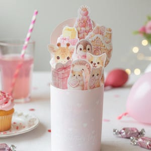 Könnte beinhalten: Eine rosa Geschenkbox aus Papier mit herzförmigen Ausschnitten, gefüllt mit Papierauschnitten zum Thema Tiere, darunter eine Giraffe, ein Pinguin und ein Igel. Ein Cupcake mit rosa Zuckerguss, ein Glas rosa Getränk und rosa Luftballons sind ebenfalls im Bild.