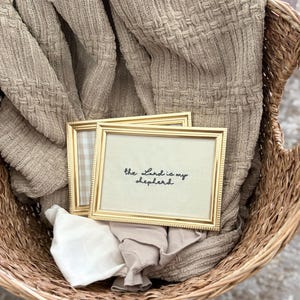 Puede incluir: Dos marcos de fotos rectangulares de tono dorado con un borde estriado. El marco frontal contiene una inserción de color crema con el texto bordado en negro "the Lord is my shepherd". El marco trasero muestra un patrón de cuadros beige claro y blanco.