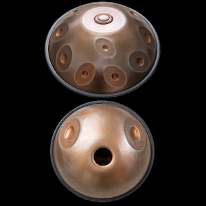 May include: A copper-colored steel handpan drum shown from two angles. The top view displays a central dome surrounded by eight circular tone fields. The bottom view shows a smooth surface with a central circular sound port and two tone fields.