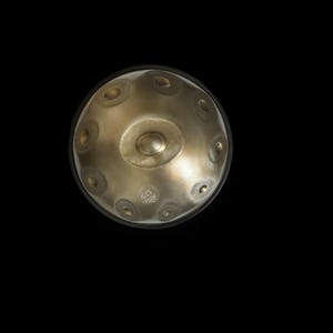 May include: A metallic gold-toned handpan percussion instrument with a smooth, rounded dome surface. It features a central raised tone field surrounded by eight circular dimpled tone fields arranged in a radial pattern, with a small logo.