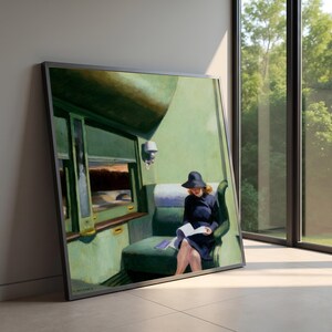 Puede incluir: Una impresión enmarcada de una pintura que representa a una persona con un vestido azul oscuro y un sombrero de ala ancha a juego, sentada en un asiento de tren verde mientras lee un libro blanco. El interior muestra paredes verdes y un aplique.