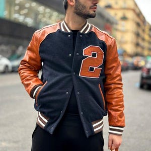 Puede incluir: Una chaqueta universitaria con cuerpo de mezcla de lana azul marino oscuro y mangas de cuero color canela. La chaqueta tiene un gran parche color canela con el número 2 en el pecho izquierdo, cierre frontal con botones a presión y ribetes a rayas.
