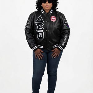 May include: A black leather varsity jacket featuring white striped ribbed cuffs and collar, Greek letters on the chest, and various embroidered patches and text on the sleeves. The jacket is worn with dark blue denim jeans and black sneakers.
