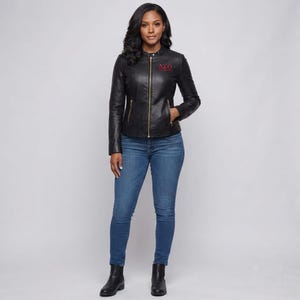 May include: A black leather jacket with a gold front zip, two gold zipped side pockets, and a small red embroidered Greek letter design on the chest. The jacket is worn with blue denim jeans and black leather ankle boots.