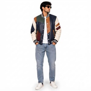 May include: A color-blocked varsity jacket featuring panels of navy blue, brown, dark green, and cream leather. The jacket has a white ribbed collar with black stripes, white ribbed cuffs, a white t-shirt, light blue denim jeans, and white sneakers.