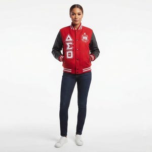 May include: A red varsity jacket with black sleeves, featuring white striped ribbed cuffs and collar. The front displays white Greek letters and a circular embroidered crest on the chest, paired with dark blue denim jeans and white trainers.