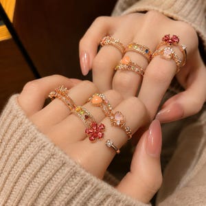 Op de afbeelding: Een collectie roségoudkleurige ringen gedragen aan meerdere vingers. De ringen hebben diverse ontwerpen, waaronder kleine heldere kristallen, roze en oranje hartvormige stenen en bloemvormige clusters van roze druppelvormige kristallen.