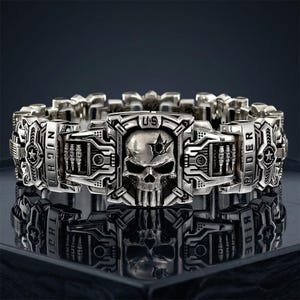 May include: A silver-toned metal bracelet featuring a central skull emblem with a bullet hole detail on the forehead. The links are engraved with the text NIGHT RIDER and US, alongside star motifs and mechanical patterns with a polished finish.
