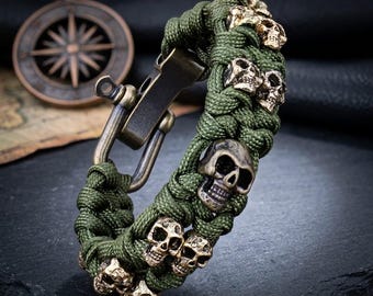 May include: A bracelet crafted from olive green paracord woven in a thick pattern, featuring multiple small metallic skull beads in a brass finish. The bracelet is secured with a large, dark bronze-toned metal shackle clasp mechanism.
