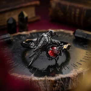 May include: A black metal ring featuring a detailed bird figure perched on a branch-like band. The bird is positioned next to a circular, deep red gemstone held within a dark, textured setting that mimics twisted vines or organic growth.