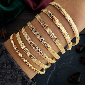 Może przedstawiać: Zestaw siedmiu złotych metalowych bransoletek typu bangle o różnych fakturach i wzorach, w tym skręcony wzór liny, motywy gwiazd, geometryczne wzory kratki i grawerunki w kształcie serca, noszone razem na nadgarstku.