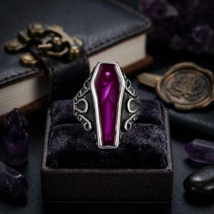 Puede incluir: Un anillo de tono plateado con una pieza central en forma de ataúd con una incrustación translúcida de color púrpura intenso. La banda de metal muestra intrincados patrones de volutas en los lados, proporcionando un aspecto detallado.