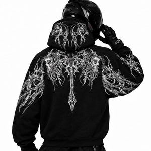 May include: A black hooded sweatshirt featuring an intricate, white, symmetrical, thorn-like graphic print that extends across the back, shoulders, and hood. The garment is worn with a black motorcycle helmet and black gloves.