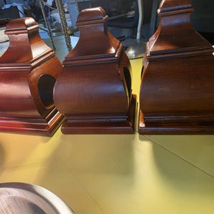 Puede incluir: Tres remates de madera de color marrón oscuro con base cuadrada y cuerpo curvo que presenta un recorte circular en la parte frontal. Cada pieza tiene una sección superior escalonada. Se ve una cinta métrica amarilla a la derecha con la marca de 5 pulgadas.