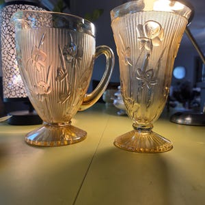 May include: A golden amber glass pitcher and a matching tall glass tumbler, both featuring a vertical ribbed texture and a raised floral pattern. Each piece stands on a circular pedestal base with a flared rim and a translucent finish.