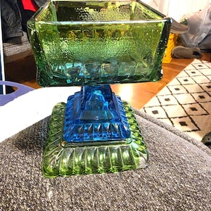May include: A square glass pedestal bowl featuring a textured green upper basin and a vibrant blue mid-section, supported by a square green base with a decorative scalloped edge. The glass displays a multi-colored design with distinct geometric patterns.
