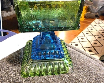 Puede incluir: Un cuenco cuadrado de cristal sobre pedestal con un recipiente superior verde texturizado y una sección central azul vibrante, apoyado sobre una base cuadrada verde con un borde festoneado decorativo. El cristal muestra un diseño multicolor.