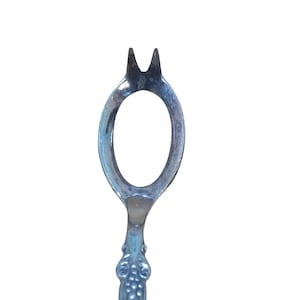 May include: A silver-toned metal serving fork featuring a unique oval-shaped head with two pointed tines at the top. The handle displays a decorative embossed pattern with small circular details, showing a metallic finish and signs of age.