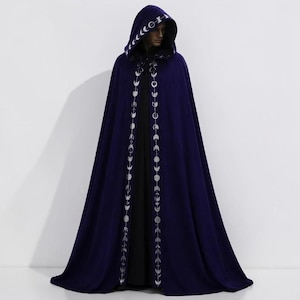 May include: A floor-length deep purple hooded cloak featuring a silver moon phase pattern embroidered along the front edges and the hood trim. The garment is worn over a solid black base layer, creating a long, flowing silhouette with a pointed hood.