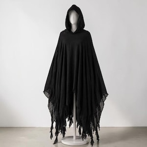 May include: A solid black hooded poncho featuring a lightweight, semi-sheer fabric with a draped, flowing silhouette. The garment includes a pointed hood and an uneven, jagged hemline finished with long, thin fabric fringe strips throughout.