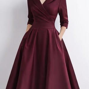 May include: A deep burgundy midi dress featuring a wide shawl collar, three-quarter length sleeves, and a fitted bodice with a wrap-style front. The skirt is pleated at the waist, creating a full silhouette, and includes side pockets.