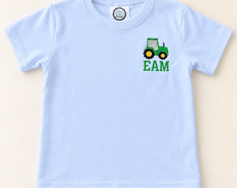 Könnte beinhalten: Ein hellblaues Kurzarm-T-Shirt mit einer gestickten grünen Traktorgrafik auf der linken Brustseite. Unter dem Traktor sind die Buchstaben EAM in grünem Garn gestickt. Das T-Shirt hat einen Rundhalsausschnitt und ein Markenetikett am Kragen.