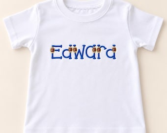 Könnte beinhalten: Ein weißes kurzärmeliges T-Shirt mit dem Namen Edward, der in blauem Garn auf die Brust gestickt ist. Kleine braun-weiße Football-Symbole sind in den Schriftzug integriert und ersetzen die vertikalen Striche der Buchstaben.