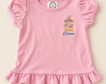 Könnte beinhalten: Ein hellrosa Kurzarmshirt mit gerafften Puffärmeln und einem gerüschten Saum. Auf der Brust befindet sich eine gestickte Karussell-Grafik in Grün, Pink und Gelb, darunter ist der Name Carmi in blauem Garn aufgestickt.