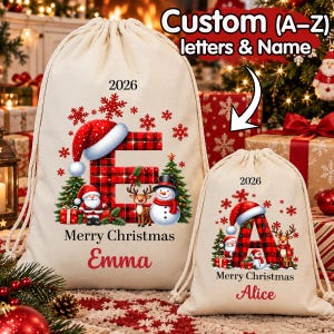 Pode incluir: Dois sacos de lona com cordão em tom off-white, com uma letra em xadrez vermelho e preto, um gorro de Pai Natal, um boneco de neve, uma rena e flocos de neve vermelhos. Os sacos exibem o ano 2026, o texto Merry Christmas e os nomes Emma e Alice.