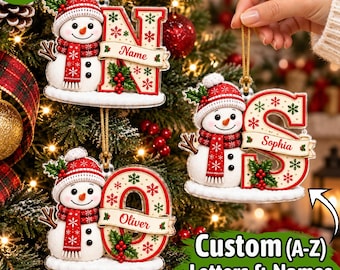 May include: Three Christmas ornaments featuring a snowman wearing a red hat and scarf next to a large letter. Each letter is decorated with red and green snowflakes, holly berries, and a banner displaying a name. The ornaments hang by gold cords.