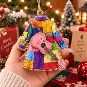 Könnte beinhalten: Ein transparenter Acryl-Weihnachtsbaumschmuck in Form eines Patchwork-Mantels mit buntem Blumen- und Geometriemuster. Eine kleine rosa Akustikgitarre mit Schmetterlingsgrafik ist vorne befestigt. Der Schmuck verfügt über eine goldene Aufhängeschlaufe.