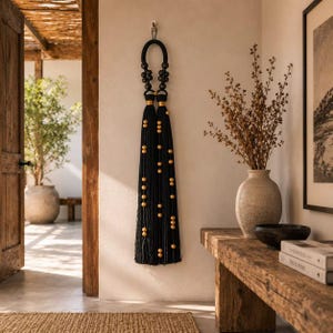 May include: A long decorative wall hanging featuring a thick black cord loop at the top, adorned with clusters of black wooden beads. Two long black tassels hang down, accented with gold-coloured beads and gold-toned metal ferrules at the top.