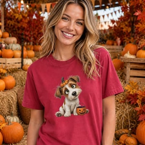 May include: A maroon short-sleeve t-shirt featuring a graphic of a small, light brown and white puppy sitting down. The puppy has a tiny pumpkin on its head and is holding a small orange jack-o'-lantern bucket in its front paws.