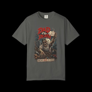 May include: A charcoal gray t-shirt featuring a graphic of a zombie rising from a grave, wearing a baseball cap and sunglasses, holding a coffee mug. The text reads DAD TIL DEATH and SOME DADS NEVER STAY BURIED, with a teddy bear and toy car nearby.