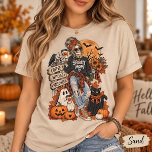 May include: A sand-coloured t-shirt featuring a graphic print of a skeleton character wearing a headband, heart-shaped sunglasses, and a plaid shirt tied around the waist. The design includes the text "SPOOKY MOM" and "COFFEE WITCHES HALLOWEEN" alongside illustrations of a black cat, a ghost, pumpkins, sunflowers, a raven, and a full moon.