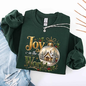 May include: A dark green crewneck sweatshirt featuring a graphic print with the text Joy in the World. The design includes a gold-toned Christmas ornament containing a snowy cabin scene with pine trees, surrounded by holly berries and gold snowflakes.