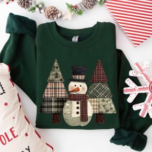 May include: A dark forest green crewneck sweatshirt featuring a central applique design of a snowman wearing a black top hat and a plaid scarf, flanked by two stylized Christmas trees made of various plaid and patterned fabric patches.