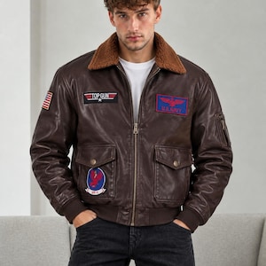 May include: A dark brown leather bomber jacket featuring a brown sherpa collar, a front zipper, and ribbed cuffs. The jacket displays various patches, including a Top Gun logo, a U.S. Navy emblem, a VF-1 patch, and a small American flag on the sleeve.
