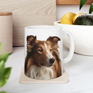 May include: A white ceramic mug featuring a detailed, realistic portrait of a sable and white Shetland Sheepdog. The dog has pointed ears, a long snout, and a fluffy white chest. The mug has a standard handle and sits on a square coaster.