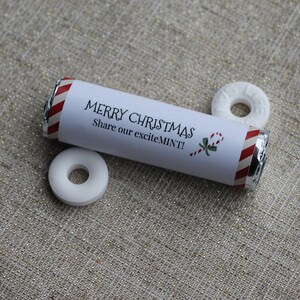 Op de afbeelding: Een witte en rood gestreepte tube met muntjes met een wit label waarop staat "Merry Christmas! Share our excite MINT!" met een afbeelding van een snoepstok en een hulsttak. Drie witte ronde muntjes liggen naast de tube.
