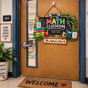 Puede incluir: Un letrero de madera para puerta de aula con diseño de pizarra, el texto MATH CLASSROOM, una pila de libros coloridos, una manzana roja, una calculadora y una placa personalizada Mrs. Anderson's Class, rematado con un lazo de cuadros.