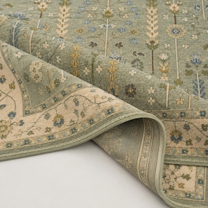 May include: A sage green area rug featuring a repeating vertical floral pattern in shades of blue, cream, and gold. The rug has a wide cream-colored border with a delicate floral vine motif and a finished edge, shown folded to reveal the underside.